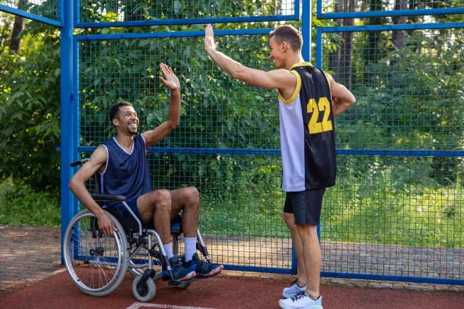 Disabled man in wheelchair with support worker high fiving on outdoor basketball court