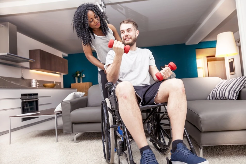 Support worker with disabled man in wheelchair in living room lifting weights